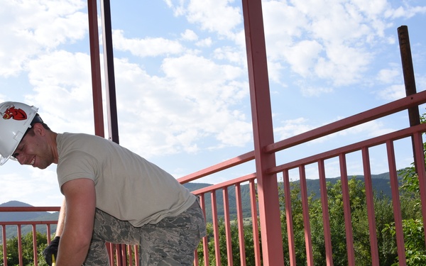 A military service member wearing camouflage uniform (ABU pattern) and a white hard hat is kneeling on a concrete surface, performing construction or demolition work on what appears to be an elevated platform or bridge. The worker is using a hand tool, likely a hammer or pry bar, to work on the concrete deck. The structure features red metal railings and is surrounded by green vegetation and mountains in the background.