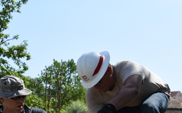 Two workers are constructing a concrete block wall outdoors in what appears to be a sunny day. One worker wears a military-style camouflage hat and dark clothing, while the other wears a white hard hat with a red stripe and light-colored clothing. They appear to be laying and aligning cinder blocks using tools and a guide string.