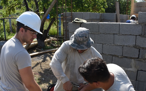 Four workers in military-style uniforms are collaborating on a construction site, focused on repairing or assembling a green wheelbarrow. They are surrounded by cinder block walls under construction, building materials, and another wheelbarrow nearby. The setting appears to be an outdoor humanitarian or military engineering project.