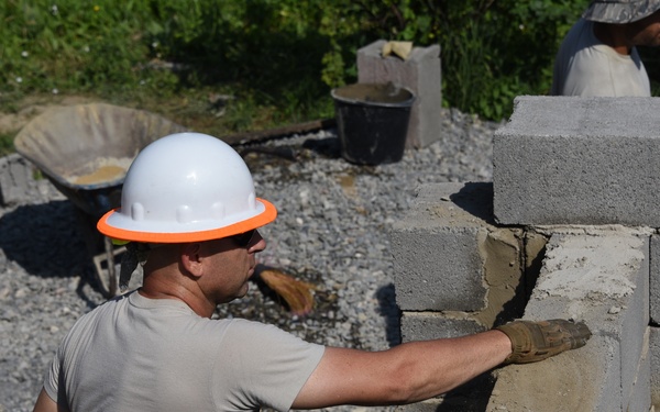A construction worker wearing a white hard hat with an orange brim is laying concrete masonry blocks, applying mortar to build a wall. At least two additional workers are visible in the background, also engaged in construction activities. The setting appears to be an outdoor building site with multiple partially constructed concrete block walls.