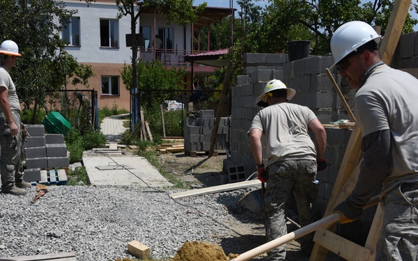 A construction site showing three workers in military-style camouflage uniforms and hard hats actively building a structure with concrete blocks. One worker in the foreground is shoveling sand into a wheelbarrow, a second worker is handling lumber near a partially constructed cinder block wall, and a third worker stands in the background observing stacked blocks. The setting appears to be a residential area with existing buildings visible in the background.