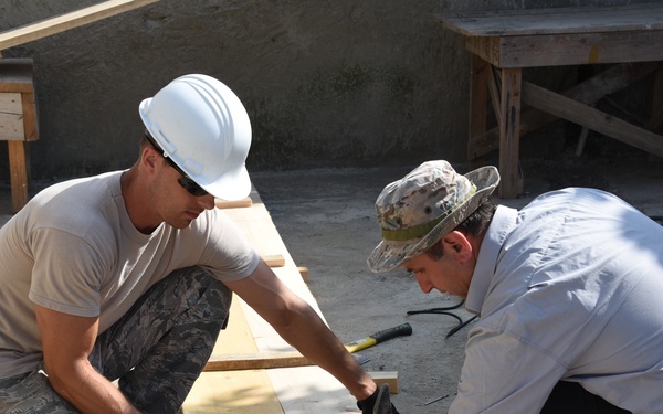 Two workers are collaborating on a carpentry task at an outdoor construction site, working with wooden planks on the ground. One worker wears US military-style camouflage pants and a white hard hat, while the other wears civilian clothing and a camouflage cap. They appear to be nailing or assembling wooden boards together.