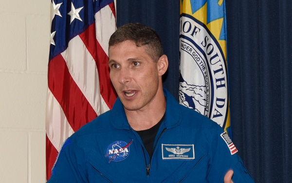 A NASA astronaut identified by name tag as Mike Hopkins is speaking or presenting while wearing a blue NASA flight suit. He is gesturing with both hands raised, standing in front of an American flag and what appears to be a South Dakota state flag. Multiple mission patches and insignia are visible on his flight suit.