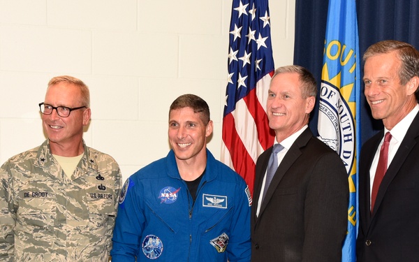 Four men are posing together for a formal group photo in front of an American flag and what appears to be a South Dakota state flag. The group includes a U.S. Air Force officer in camouflage uniform, a NASA astronaut in a blue flight suit, and two men in civilian business attire. The setting appears to be an official or ceremonial event.