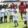 An outdoor youth football camp hosted by Andre Roberts is taking place on a grass field. A tall adult wearing a red 'Andre Roberts' branded shirt, bucket hat, and sunglasses supervises several young children in white camp t-shirts. Two children appear to be actively running a drill while others wait in line.