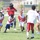 An outdoor youth football camp is taking place on a grass field, with an adult instructor wearing a red 'Andre Roberts' shirt and black bucket hat holding a football while engaging with a group of children. The kids are wearing red and white camp jerseys and appear to be participating in a flag football or skills drill activity. A modern brick building is visible in the background, suggesting a school or community facility setting.