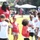 A tall adult male wearing a red 'Andre Roberts' branded shirt and black bucket hat holds a football while interacting with a group of young children on a grass field. The event appears to be a youth football camp or clinic organized by Andre Roberts, as evidenced by branded shirts and red mesh vests worn by participants. The outdoor event takes place on a sunny day with canopy tents and spectators visible in the background.