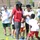 An outdoor youth football camp is taking place on a grass field, led by an adult instructor wearing a red shirt labeled 'Andre Roberts' and holding a football. Multiple children in white camp t-shirts are participating in drills, with another adult coach visible on the left side. The setting appears to be near a school or community facility on a sunny day.