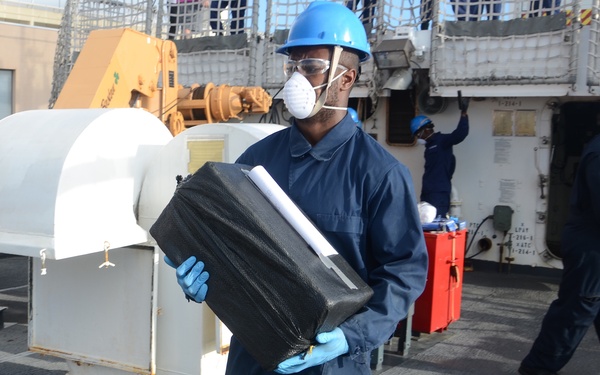 Personnel in blue coveralls and blue hard hats are working on the deck of what appears to be a military or coast guard vessel. The foreground figure is carrying a large black case while wearing PPE including a respirator mask, safety glasses, and blue nitrile gloves. Additional crew members are visible in the background performing tasks on deck.