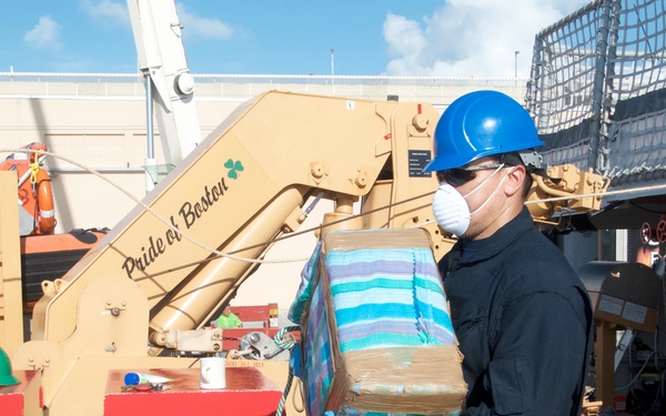 A person wearing a blue hard hat, respirator mask, and blue latex gloves is carrying a wrapped package or bale aboard what appears to be a military or coast guard vessel. The equipment visible includes a tan-colored crane or davit arm labeled 'Pride of Boston' and red equipment containers marked 'WARNING F-76' and 'BNTNG F-76'. The scene suggests an official interdiction or cargo transfer operation on a ship's deck.