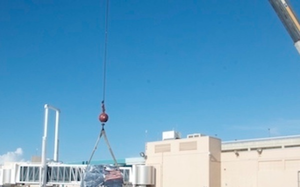 A U.S. Coast Guard rigid inflatable boat (RIB) is being lifted or positioned by a crane at what appears to be a port or military facility. Workers in safety gear are visible on the dock overseeing the operation. An American flag is prominently displayed on the left side of the image.