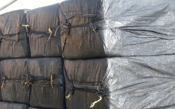The image shows a large pallet stacked with multiple tightly wrapped black bales secured with rope and twine, consistent with contraband seizure documentation commonly seen in law enforcement or coast guard operations. The bales are uniformly packaged in dark woven plastic material and stacked in a grid pattern approximately four rows high. The setting appears to be on a vessel or dock based on the wooden pallet and rope visible in the background.