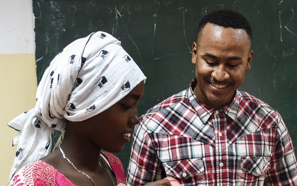 Two individuals are standing in front of a green chalkboard in what appears to be a classroom setting. A woman wearing a white patterned headscarf and pink floral dress is holding a dental model (jaw/teeth model) and a toothbrush, while a man in a red plaid shirt smiles and observes. The scene suggests a dental hygiene demonstration or educational session.