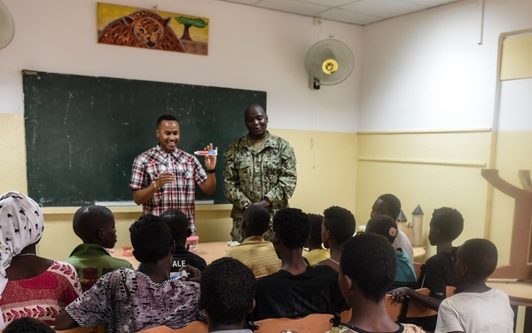 The image shows a classroom setting where two adults, one in a plaid shirt and one in military uniform, are presenting to a group of children seated in wooden chairs. A third adult in civilian clothing stands to the left observing. The setting appears to be a school in an African country, with a green chalkboard and a wildlife painting visible on the wall.