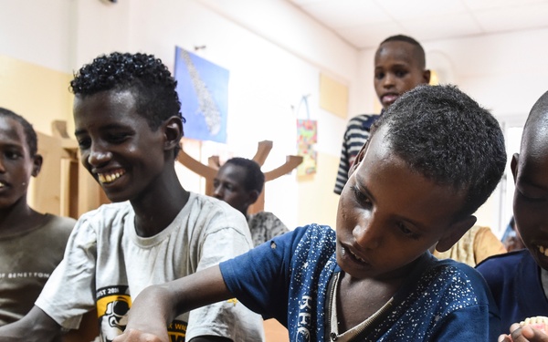 A group of young boys are participating in a dental hygiene education session in what appears to be a classroom or community health setting. The children are actively engaged with dental model props, practicing toothbrushing techniques on plastic jaw models. The atmosphere appears lively and interactive, with the children showing visible enthusiasm.