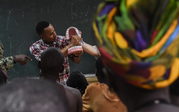 A dental health education session is taking place in a classroom setting. An instructor is holding up a large dental model (jaw/teeth model) to demonstrate oral hygiene techniques to an audience of seated attendees. A person in military camouflage uniform with a visible US flag patch is also present at the front of the room.