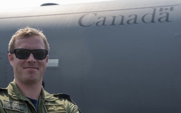 A Canadian military aviator in an olive green flight suit stands with arms crossed in front of a large grey Canadian Armed Forces aircraft. The individual is wearing sunglasses and a wristwatch, and displays a name tag and wing insignia on their flight suit. The aircraft bears the word 'Canada' with a maple leaf emblem on its fuselage.