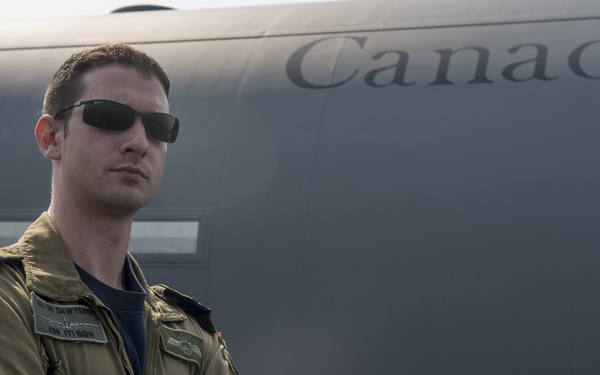 A military aviator in an olive/khaki flight suit stands with arms crossed in front of a Canadian Armed Forces aircraft. The individual wears sunglasses and displays a unit patch on the left shoulder, along with name tags on the chest. The aircraft behind him bears the word 'Canada' in large lettering on its fuselage.
