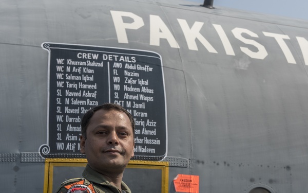 A Pakistan Air Force aviator in olive green flight suit stands with arms crossed in front of a military aircraft fuselage. The aircraft bears the word 'PAKISTAN' in large white letters and displays a 'Crew Details' placard listing multiple crew members' names and ranks. A Squadron 21 Pakistan Air Force patch (featuring Pegasus) is visible on the aircraft, along with multiple patches on the aviator's flight suit.