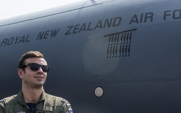 A Royal New Zealand Air Force personnel member stands in front of a large military aircraft bearing the inscription 'ROYAL NEW ZEALAND AIR FORCE'. The individual is wearing an olive green flight suit with patches and a name tag reading 'KENDALL DOOLEY', and is posing with arms crossed while wearing sunglasses. The image appears to be an official or promotional military photograph taken on an airfield.