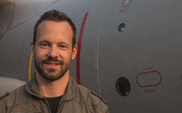 A military pilot in an olive/khaki flight suit stands in front of a large gray military aircraft. His name tag reads 'VAN DYCK G.' with the callsign 'GILLES' and designation 'PILOT', and he wears multiple patches including a squadron patch labeled 'TENACIY' and what appears to be a Belgian flag patch. The image is taken during golden hour lighting conditions.