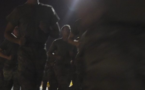 A nighttime photograph showing a group of military personnel in uniform gathered or moving on a paved surface. The image is taken at low angle with a blurred foreground figure, illuminated by artificial lighting. The dark conditions and motion blur reduce overall image clarity.