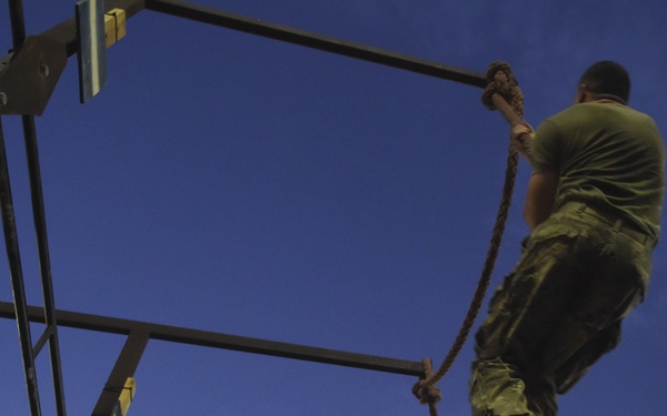 A person in military-style camouflage clothing and boots is climbing a thick rope attached to a metal frame structure, photographed from a low angle looking upward. The image is taken at dusk or dawn, with a deep blue sky in the background. This appears to be a military or fitness obstacle course training exercise.