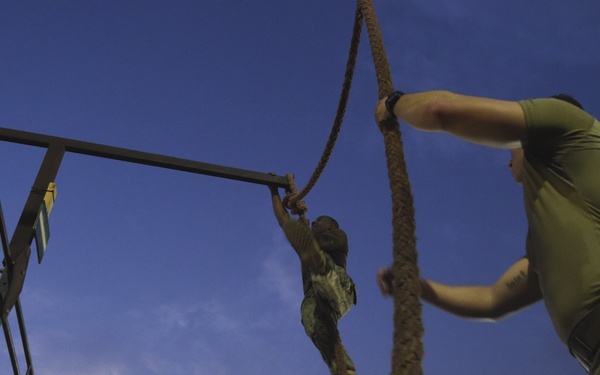 Military personnel are conducting a nighttime physical training exercise on an obstacle course. One soldier is climbing a rope suspended from a metal frame structure while others observe and assist from the ground. The scene is illuminated by stadium lighting against a twilight sky.