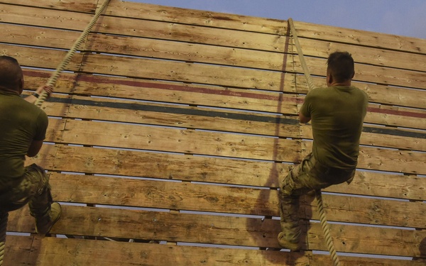 Two soldiers in military camouflage uniforms are climbing a large wooden wall obstacle using ropes, viewed from a low angle looking upward. A third soldier stands to the right holding a clipboard, appearing to observe and evaluate the performance. This appears to be a military obstacle course or physical fitness training exercise.