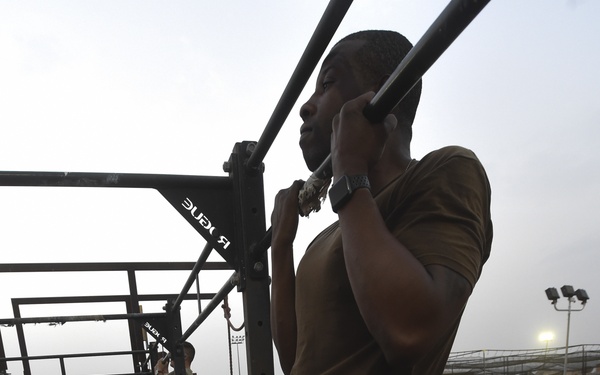 A military service member is performing pull-ups on a Rogue fitness rig while being observed by a group of fellow service members in military uniforms. The setting appears to be an outdoor military fitness area or obstacle course, likely during a physical fitness test or competition. Multiple branches of service appear to be represented based on the varying camouflage patterns visible.