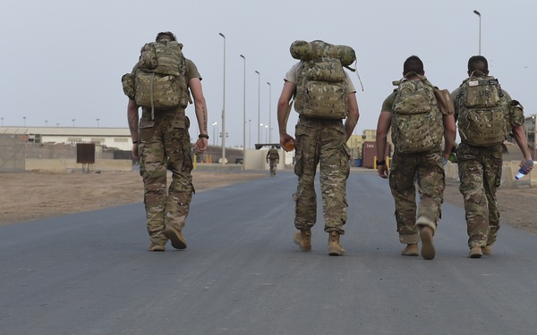 Four military personnel in camouflage uniforms and tactical backpacks are walking away from the camera down a paved road on what appears to be a forward operating base or military installation. The arid, desert environment with sandy terrain, utility poles, and military structures visible in the background suggests a location in a hot, dry region. A fifth individual can be seen further down the road in the distance.