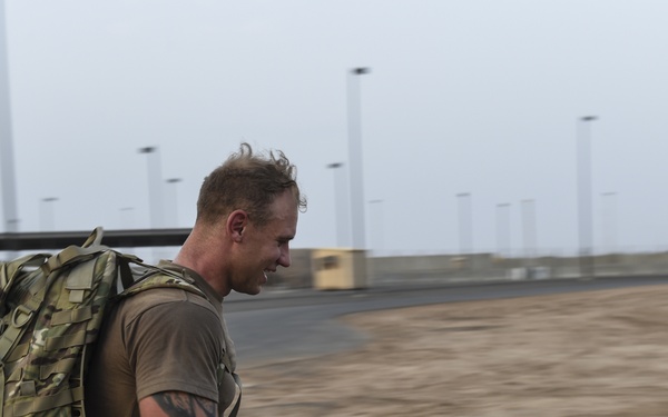 A male military service member is running or rucking (ruck marching) while carrying a heavy camouflage tactical backpack on a paved road at what appears to be a military installation. He is wearing a brown t-shirt, camouflage pants, and has a visible tattoo on his left arm. The background shows a flat, arid environment with light poles and military infrastructure.