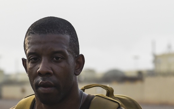 A man wearing a brown shirt and olive/tan tactical backpack is photographed outdoors in what appears to be an arid or desert environment. He is looking directly at the camera with a neutral expression. The background shows blurred infrastructure including walls, light poles, and what appears to be a facility or compound.