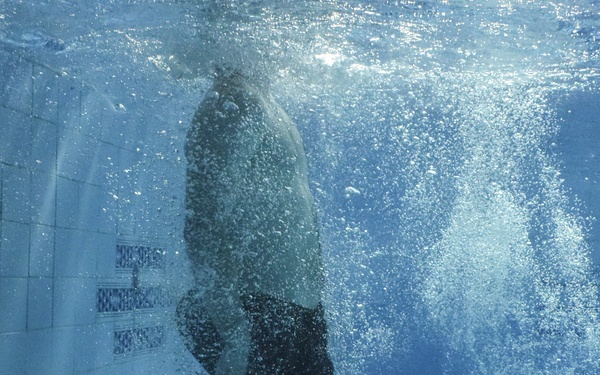 An underwater photograph taken inside an indoor swimming pool, capturing a person mid-entry or mid-dive into the water. The image shows the submerged torso and legs of an individual wearing dark swim shorts, surrounded by a dense cloud of air bubbles generated by the water entry. The pool's tiled walls and floor are clearly visible in the blue-tinted water.