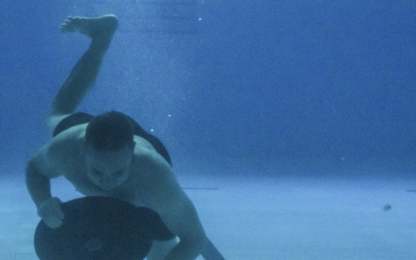 An underwater photograph taken inside a swimming pool shows a person submerged near the pool floor, holding a large dark circular disc. The swimmer appears to be engaged in an underwater sport or training exercise, possibly underwater hockey or a military/diving drill. The image is captured from a low angle, revealing the tiled pool bottom and blue water environment.