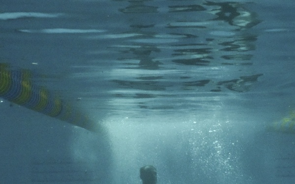 An underwater photograph taken inside an indoor swimming pool showing two individuals submerged beneath the surface. The two people appear to be interacting with a dark circular flotation ring or inner tube. Lane dividers with colorful floats are visible near the water surface.