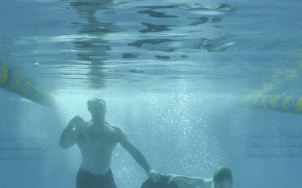 An underwater photograph taken inside a swimming pool shows two individuals submerged beneath the surface. A larger person and a smaller person (likely an adult and a child) are both underwater, appearing to compete over or share a dark circular disc or puck. The image is taken from a side/underwater perspective with lane dividers visible at the top.