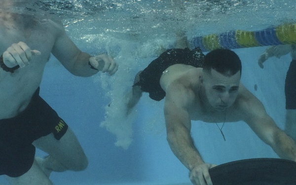 An underwater photograph taken inside an indoor swimming pool shows two shirtless men submerged and actively engaged in what appears to be an underwater hockey or disc sport. One man is pushing a large black puck/disc along the pool floor while the other moves alongside him. A third person is partially visible standing at the pool's edge above the waterline.