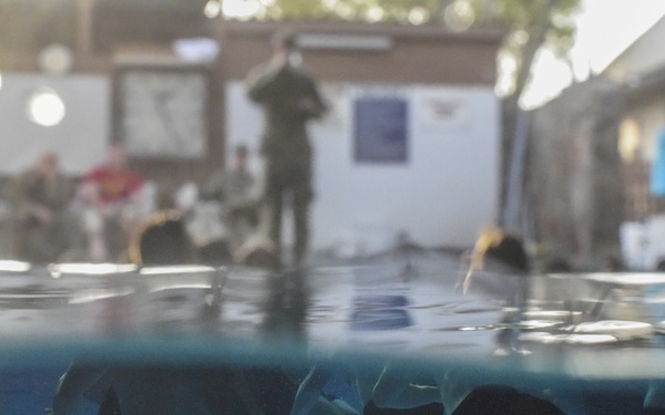 A split-level water photograph showing multiple people submerged underwater in a pool, wearing dark swim shorts. Above the waterline, several individuals are visible on the pool deck, including one standing figure in what appears to be a uniform or dark outfit. The setting appears to be an outdoor pool area with buildings and equipment visible in the background.