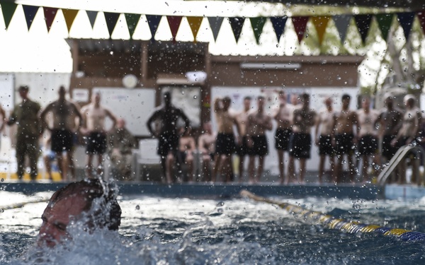 A swimmer is actively swimming in a lane pool, captured mid-stroke with significant water splash around them. A large group of spectators, many appearing to be in athletic or military-style shorts, stand poolside watching the event. Colorful pennant flags are strung overhead, suggesting an organized swimming event or competition.