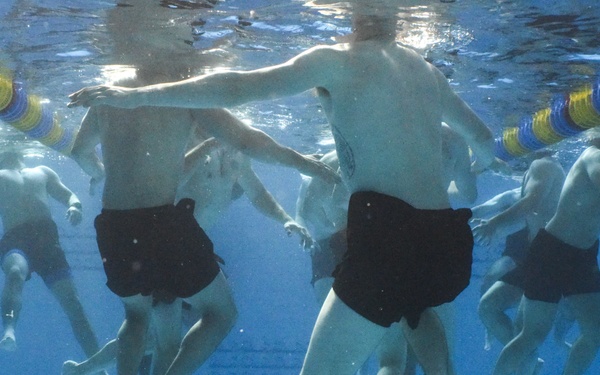 An underwater photograph taken inside a swimming pool showing multiple people treading water or performing aquatic exercises. The image is captured from below the surface, providing an upward-looking perspective of the participants. All individuals are wearing black swim shorts and are visible from approximately the waist down to their feet.