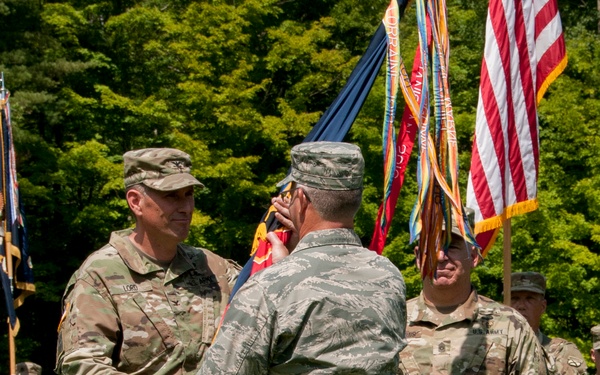 A military change of command ceremony is taking place outdoors on a grass field. Two officers are exchanging a unit guidon (flag), which is the central symbolic act of a change of command ceremony. Multiple soldiers in various uniforms are present, along with several flags including the American flag.