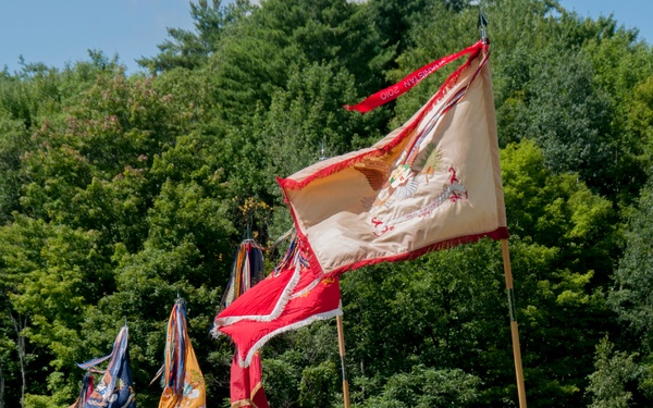A U.S. Army military ceremony is taking place on a grass field, with soldiers in OCP (Operational Camouflage Pattern) uniforms standing in formation. Multiple unit colors and guidons are being displayed, including a prominent tan/cream flag with a red streamer reading 'AFGHANISTAN 2010' and a red flag with white fringe. The ceremony appears to be a formal parade or change of command event conducted outdoors with trees and military vehicles visible in the background.