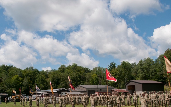 A large military formation is assembled on an open grass field, likely during a formal ceremony or parade. Multiple unit guidons and flags, including what appears to be a US flag, are visible among the formations. The setting appears to be a rural military installation or training area with farm buildings and dense tree line in the background.