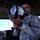 A U.S. Air Force airman is holding and operating a light gun signal device, likely in an air traffic control tower. The device, manufactured by Avionics Inc., is used to communicate with aircraft when radio communication is unavailable. The setting appears to be an elevated control tower with panoramic windows overlooking an airfield.