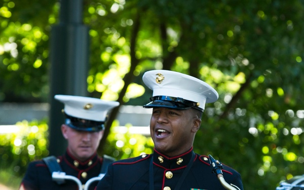 A U.S. Marine Corps band is performing outdoors, with multiple musicians in full dress blues uniforms. The central figure, a Sergeant, is playing a saxophone and appears to be singing or calling out while performing. The setting appears to be a daytime outdoor ceremony or public performance.