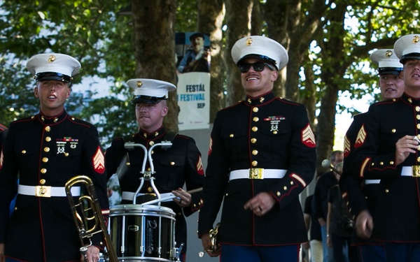 A United States Marine Corps marching band is parading in dress blues uniform along a tree-lined path. The musicians are carrying instruments including a trombone, snare drum, and trombone/trumpet, with several members displaying sergeant chevrons on their sleeves. A sign in the background references a 'Festál Festival' in September, suggesting this is a public cultural event.