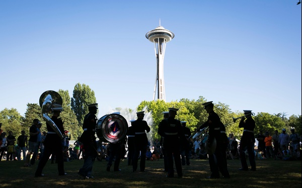 A military marching band performs in silhouette on a grassy lawn near the Seattle Space Needle on a clear, sunny day. The band members are dressed in dark uniforms with white caps and are playing various instruments including a bass drum, sousaphone, and trumpet. A crowd of spectators is visible in the background watching the performance.