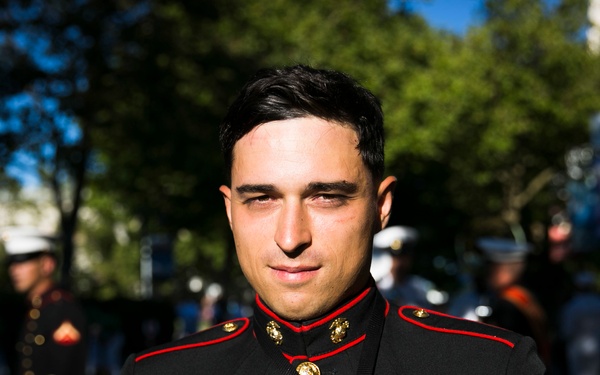A U.S. Marine Corps Sergeant is photographed in dress blues uniform at what appears to be an outdoor military parade or public event. He is wearing the distinctive black and scarlet dress uniform with gold buttons, rank chevrons, ribbons, and badges visible on his chest. Other military personnel in various uniforms are visible in the blurred background.