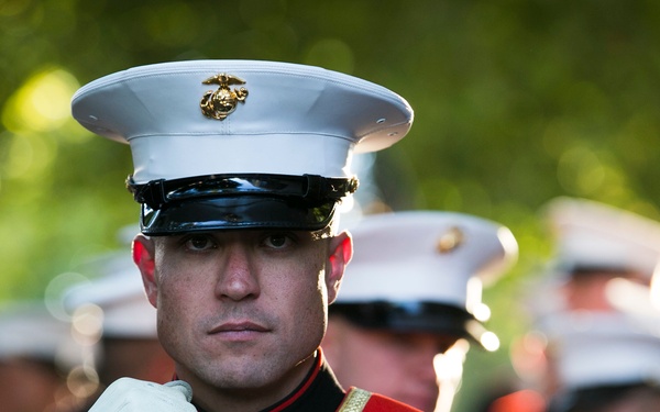 A United States Marine Corps sergeant in dress blues uniform stands at attention holding a ceremonial mace, indicating a formal military parade or ceremony. He wears a distinctive red sash with the USMC emblem and multiple service ribbons and medals. Additional Marines in dress uniform are visible in the blurred background.